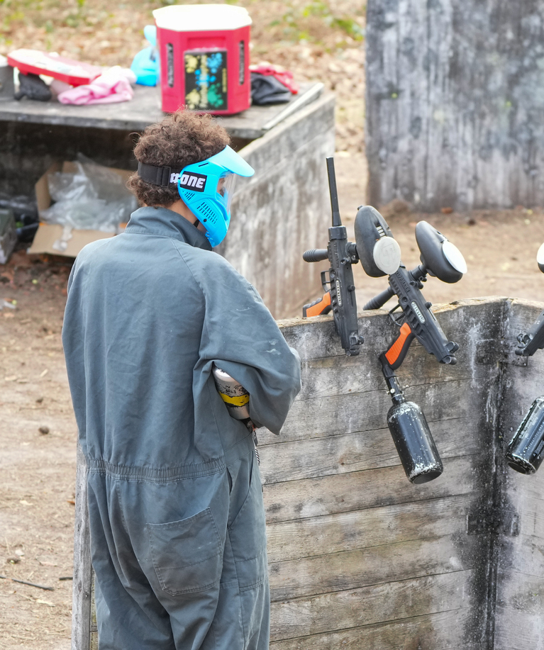 Joueur de paintball en combinaison grise et masque bleu positionné derrière un abri coloré, marqueur à la main, lors d'une fête d'anniversaire paintball à La Hulpe