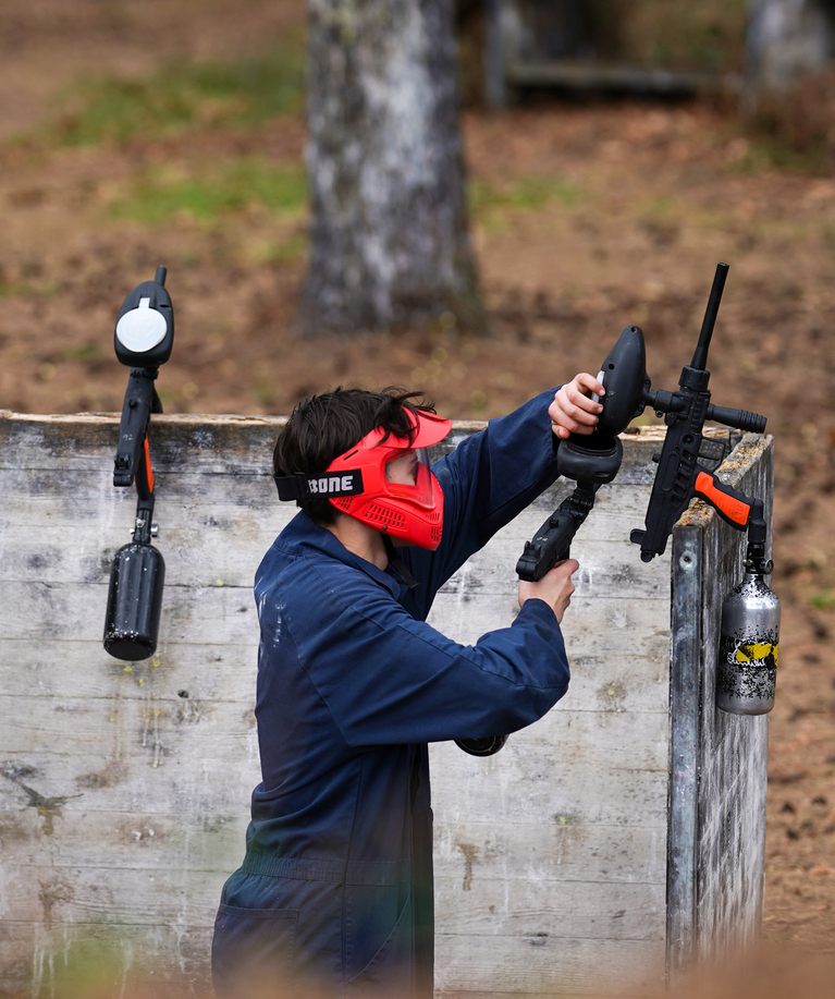 Joueur de paintball en combinaison bleue et masque rouge visant avec son marqueur par-dessus une barricade en bois lors d'une partie d'anniversaire paintball à La Hulpe
