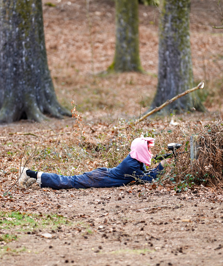 Joueur de paintball en combinaison bleue et masque rose allongé au sol dans une forêt, simulant une élimination lors d'une partie d'anniversaire paintball à Overijse