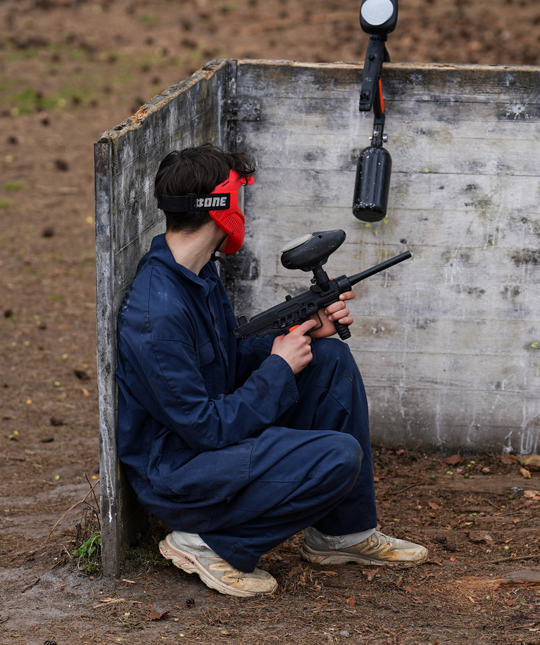 Joueur de paintball en combinaison bleue et masque rouge accroupi contre un mur, marqueur pointé vers l'avant, en attente de l'adversaire lors d'une fête d'anniversaire paintball à Rixensart