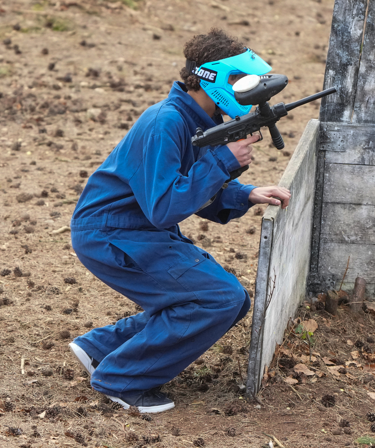 Jeune joueur de paintball en combinaison bleue et masque de protection turquoise accroupi derrière un arbre, marqueur en main, lors d'une fête d'anniversaire paintball à Tervuren