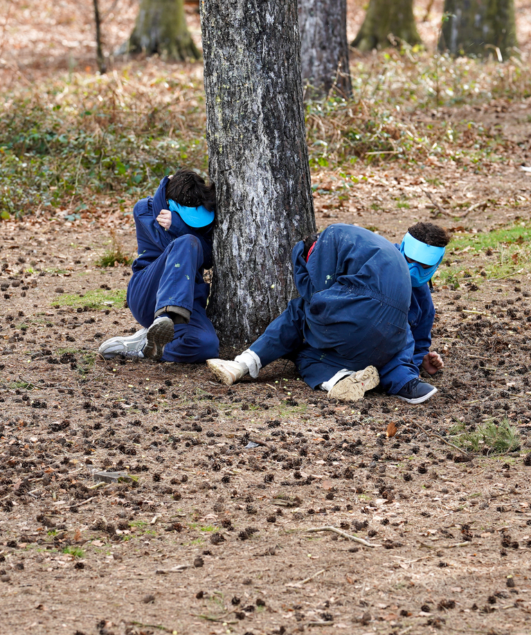 Deux jeunes joueurs de paintball en combinaisons bleues accroupis derrière un arbre en forêt, se cachant de l'adversaire lors d'une fête d'anniversaire paintball à Tervuren